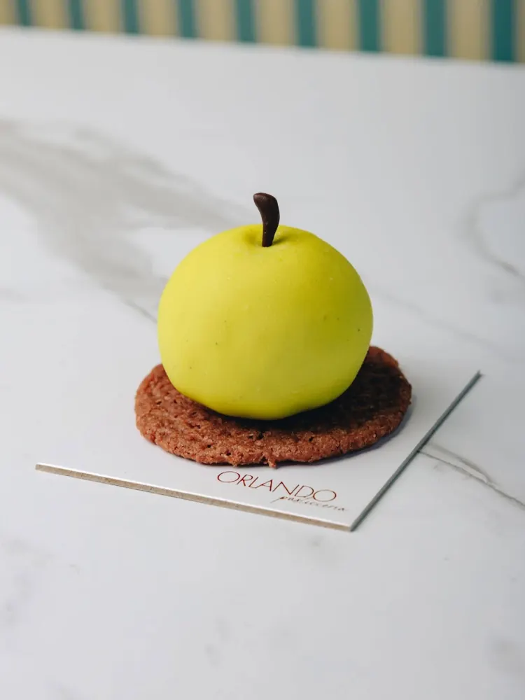 Dolce a forma di mela verde, con il picciolo di cioccolato, appoggiato su un biscotto al cacao